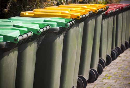 A row of large, wheeled plastic rubbish bins positioned on a paved outdoor surface, with the front of the bins facing the camera. The bins are predominantly dark grey with brightly coloured lids in green, yellow, and red, arranged in sequence along the length of the row. The surface appears to be a mixture of concrete or tarmac with small patches of moss or grass growing between the paving stones. The background shows some greenery, possibly bushes or trees, indicating an outdoor setting typical of waste collection points in residential or commercial areas. The lids are closed, and the bins are clean and well-maintained, reflecting a typical rubbish collection setup associated with local waste management services such as those provided by Office Clearance Sutton in the Sutton area.