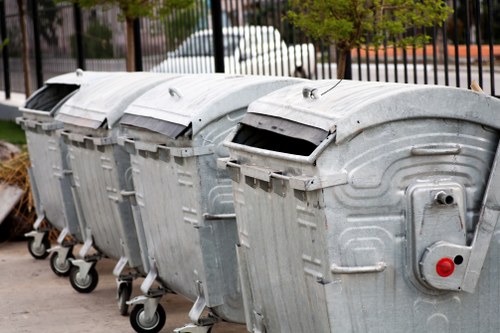 A row of five large metal wheelie bins used for rubbish collection, positioned on a paved surface outdoors. The bins are made of galvanized steel, with textured, ribbed surfaces and dark gray lids, some slightly open. Each bin has four black plastic wheels, with the front wheels visible in the foreground. The background features a black metal fence, with a white vehicle and some trees visible behind it, indicating an urban or industrial setting. The image appears to be taken during daylight hours, with natural lighting highlighting the metallic finishes of the bins and the surroundings. This scene is relevant to waste management and rubbish removal services, as provided by companies like Office Clearance Sutton in the Sutton area.