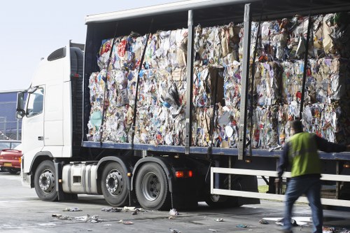 A large white commercial lorry parked on a paved area, loaded with tightly stacked bales of mixed waste materials including paper, cardboard, and plastic, visible through metal framing on the truckâ€™s side. The load appears bulky and densely packed, with some loose pieces visible on the ground near the truck. In the foreground, a person wearing a high-visibility yellow vest and dark trousers is walking alongside the vehicle, suggesting a waste collection or rubbish removal activity typical of local services in Sutton. The vehicle's cab faces left, with a clear sky overhead, and other vehicles are partially visible in the background, indicating an outdoor waste handling or disposal environment. The image captures a scene consistent with professional rubbish clearance operations by companies like Office Clearance Sutton, supporting waste management needs within the local area.