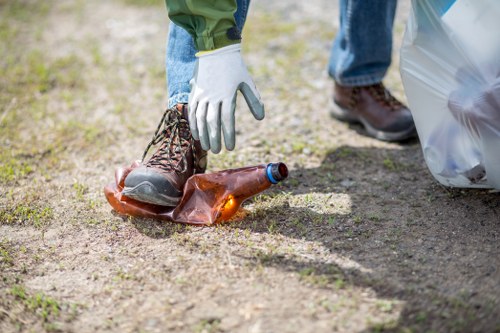 A person wearing light-colored gloves and blue jeans is seen picking up a discarded glass bottle with a dark brown color and a slim neck from a patch of dirt or gravel ground. The individual is wearing dark brown boots and is holding the bottle near its base, close to the ground. To their right, a large transparent plastic rubbish bag is partially visible, resting on the ground. The background shows an outdoor area with natural grass and soil, indicating an environment where waste has been gathered for removal. This scene illustrates the process of collecting litter, aligning with rubbish removal services such as those offered by Office Clearance Sutton, based near Sutton and relevant to local rubbish clearance activities.