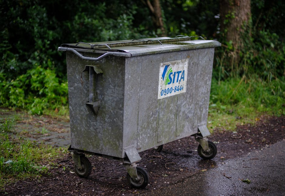 In the foreground, a row of five large, white plastic wheelie bins with closed lids are positioned on a paved surface, each equipped with handles and small wheels at the base for mobility. The bins ar