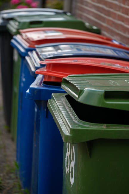 A large collection of mixed waste and rubbish is piled up on a paved sidewalk in front of a row of waste and recycling bins of various sizes and colours, including a grey bin labeled for mixed paper a