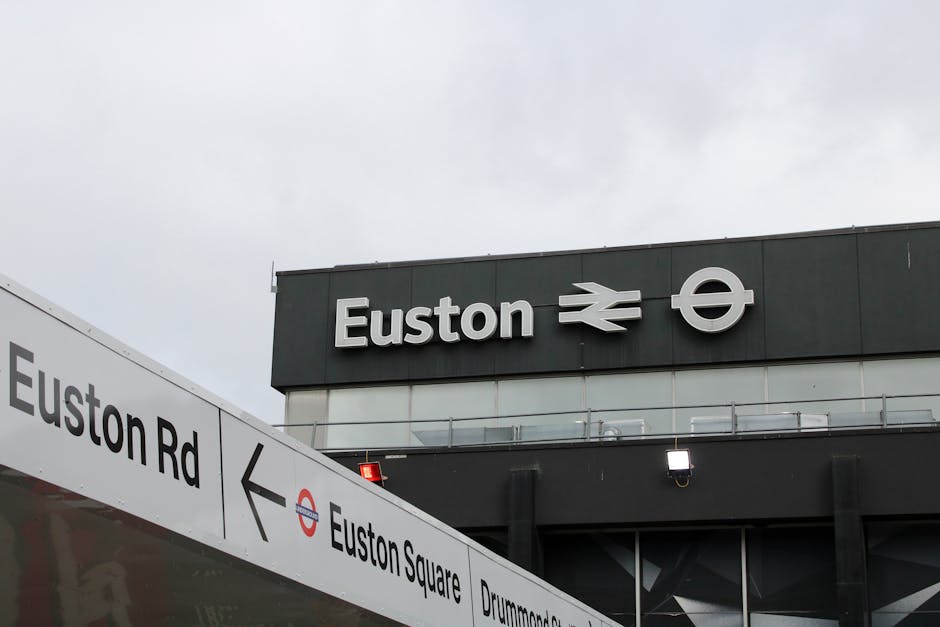 At a modern train station platform, a worker dressed in dark clothing, a beanie, and a high-visibility yellow vest stands beside a large red trolley cart filled with numerous black, gray, and orange g