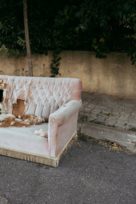 In a dingy, neglected room with dark, stained walls showing signs of decay and peeling paint, a worn, brown leather sofa with visible creases and a slightly damaged surface is positioned against the b